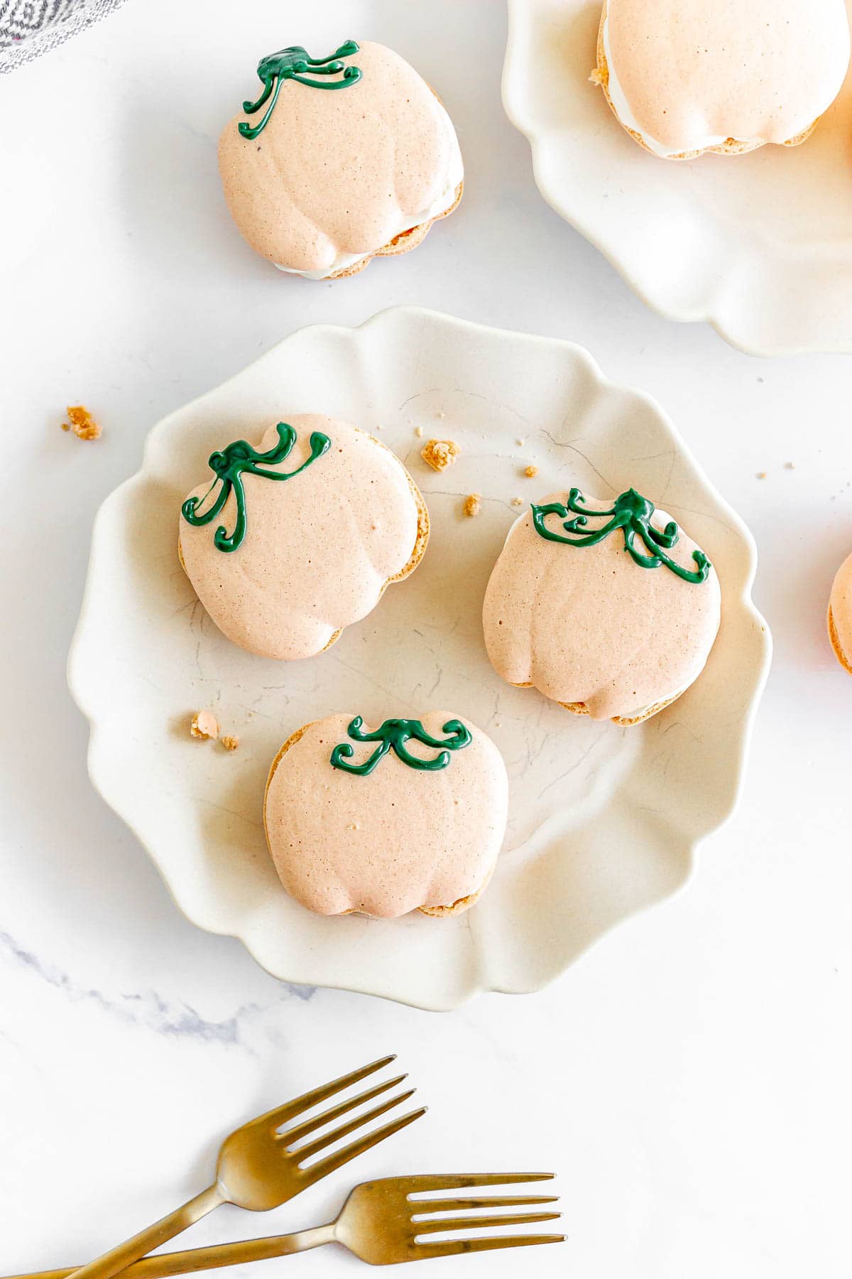Pumpkin Macarons shaped like pumpkins with green icing rest on a white plate, accompanied by two gold forks.