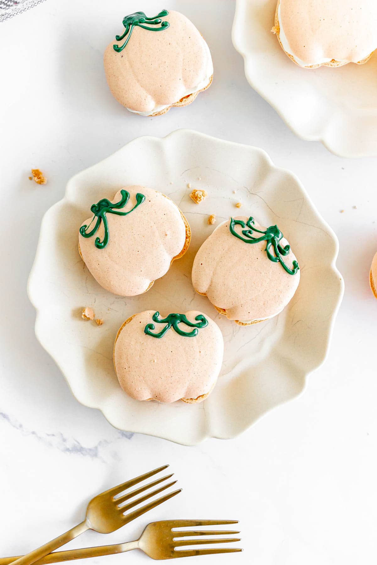 Pumpkin Macarons shaped like pumpkins, topped with green icing stems, are arranged on a white plate with gold forks beside them.