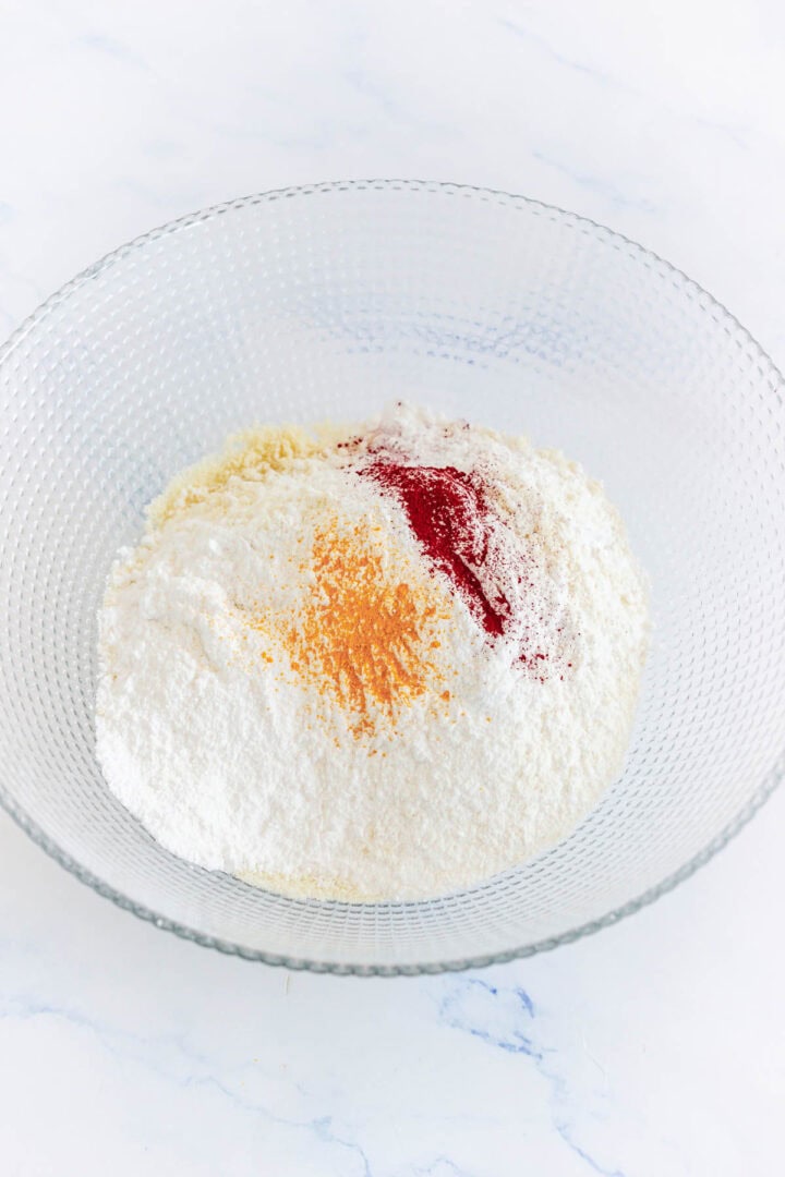 Clear glass bowl with white flour and two piles of red and yellow powdered spices, perfect for creating festive treats like Pumpkin Macarons, on a white surface.