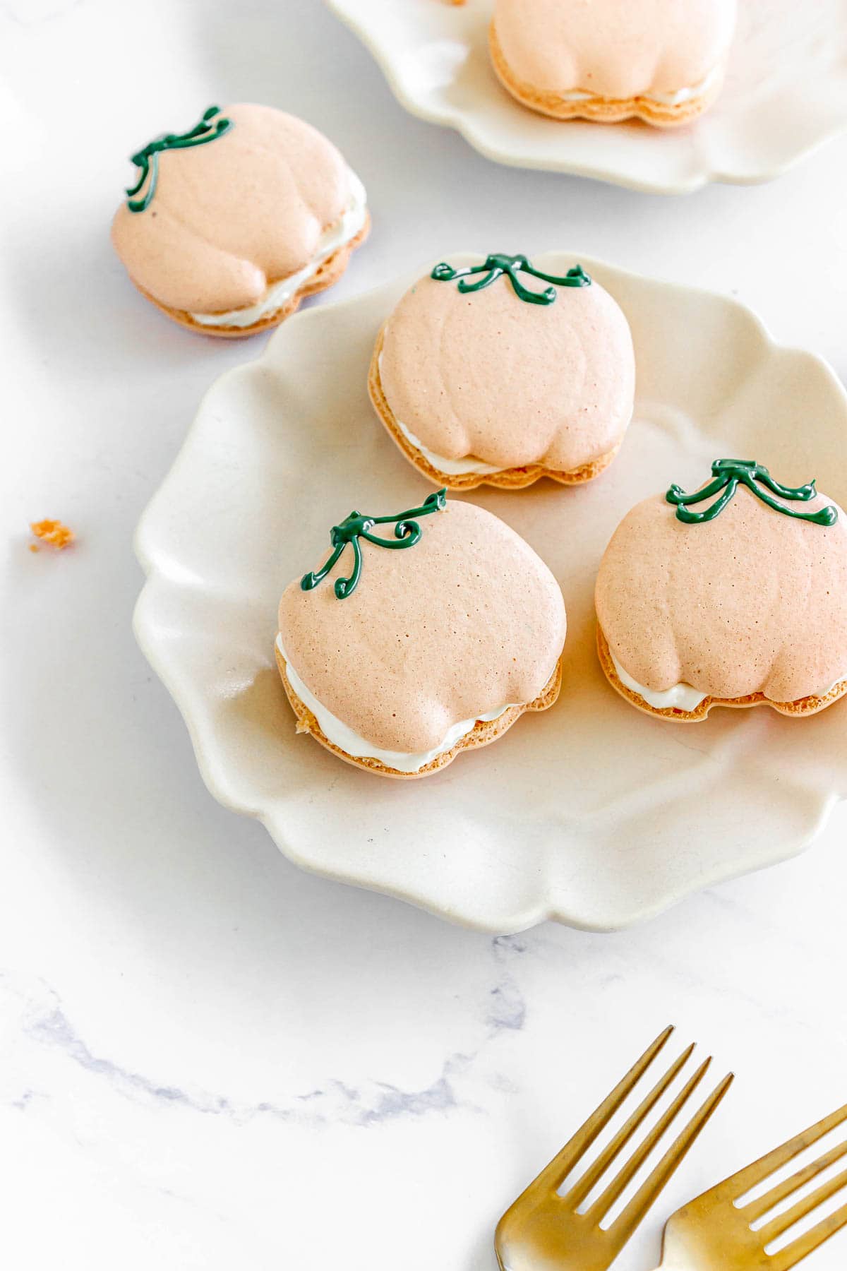 Pumpkin Macarons shaped like pumpkins with white filling rest on a white plate, accompanied by two gold forks.