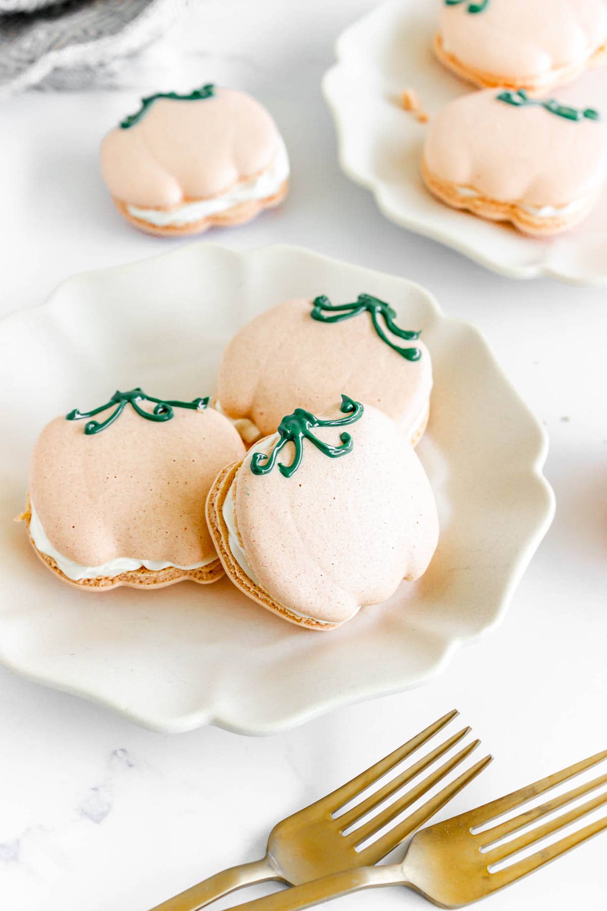 Pumpkin Macarons with cream filling are shaped like pumpkins and topped with green icing stems, arranged beautifully on a white plate.