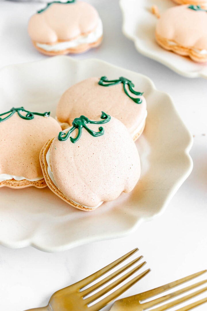 Pumpkin Macarons shaped like pumpkins with green icing stems rest on a white plate, with two forks in the foreground.