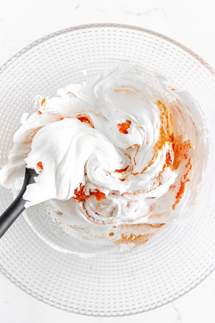 A spatula mixing white meringue with orange food coloring in a textured glass bowl, preparing the vibrant batter for Pumpkin Macarons.