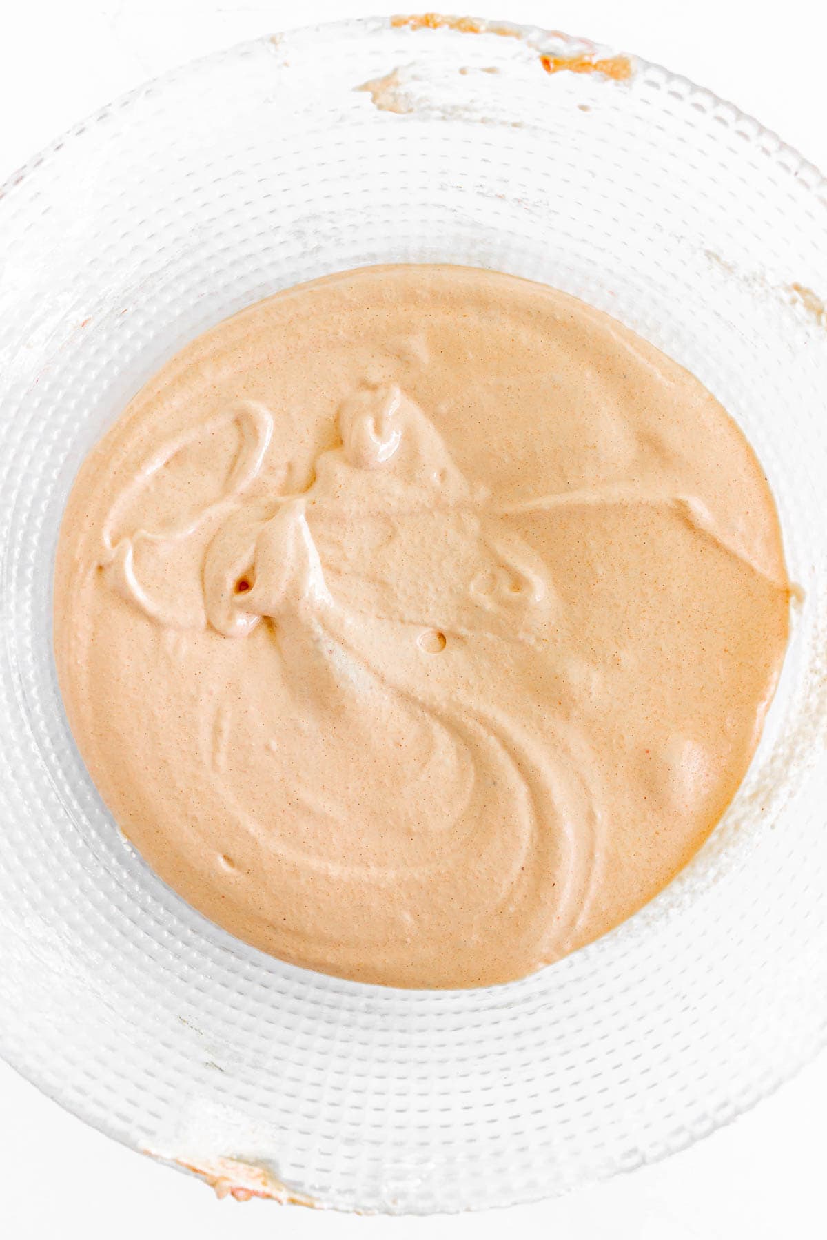A glass bowl filled with light orange, creamy batter for Pumpkin Macarons sits on a white surface.