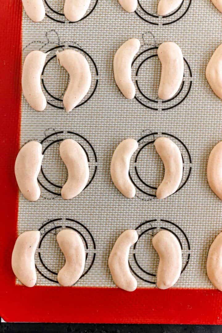 Piped crescent-shaped Pumpkin Macarons batter on a silicone baking mat with circular guides.