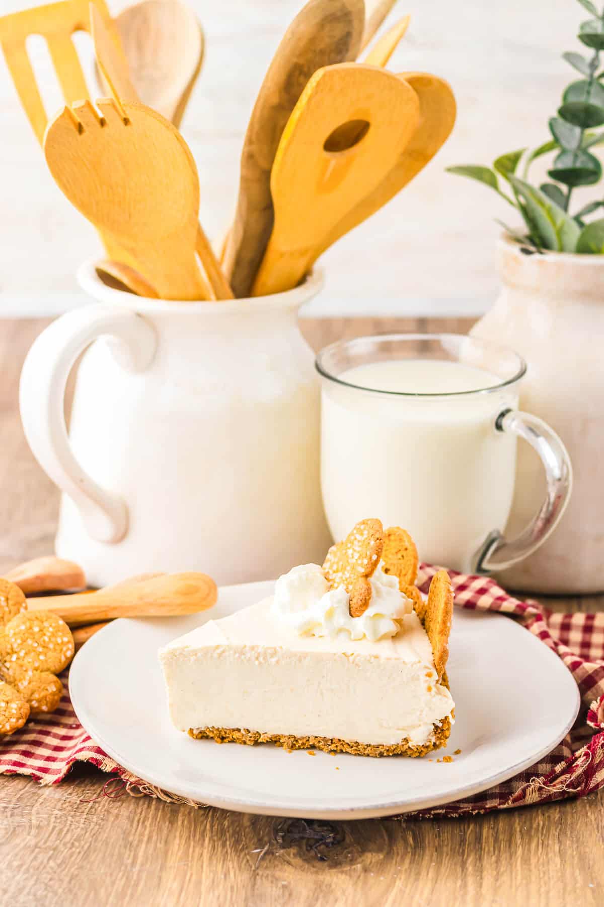 A slice of No Bake Gingerbread Cheesecake with whipped cream and cookies on a plate, next to a glass of milk and wooden utensils.
