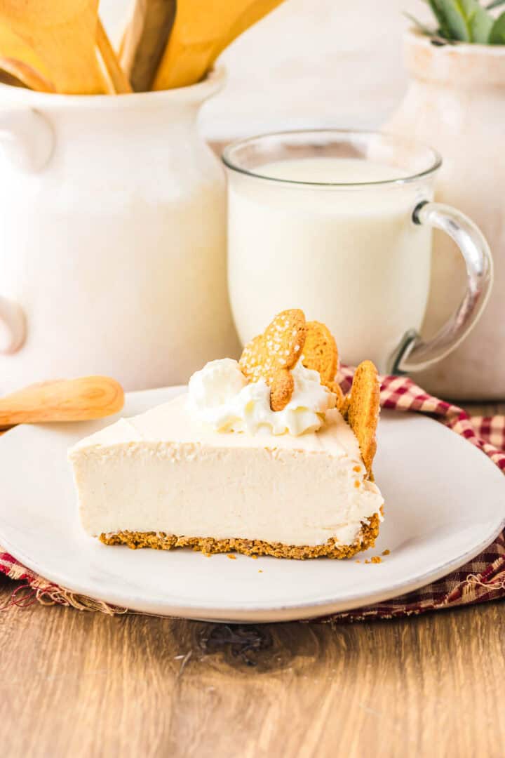 A slice of creamy No Bake Gingerbread Cheesecake with whipped cream and cookies on top, served on a white plate with milk in the background.