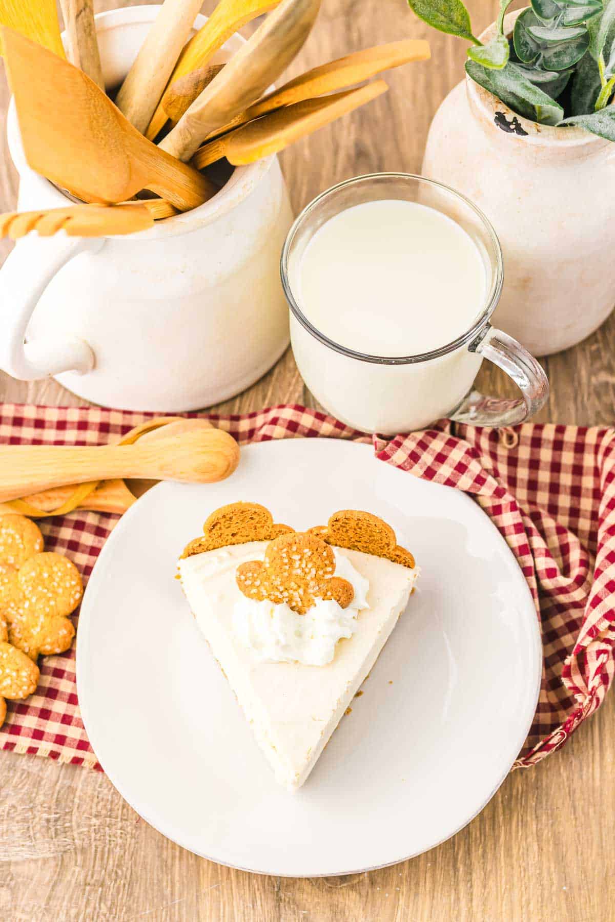 A slice of No Bake Gingerbread Cheesecake with whipped cream and cookies on a plate, next to a glass of milk on a red checkered cloth.