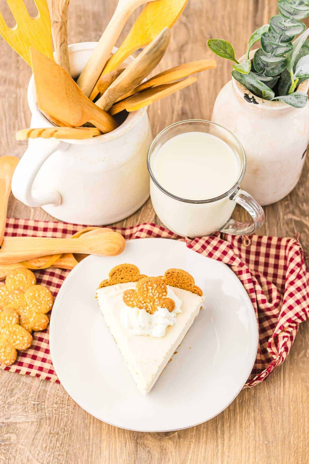 Slice of No Bake Gingerbread Cheesecake with whipped cream and gingerbread cookie, milk, and wooden utensils on a checkered cloth.