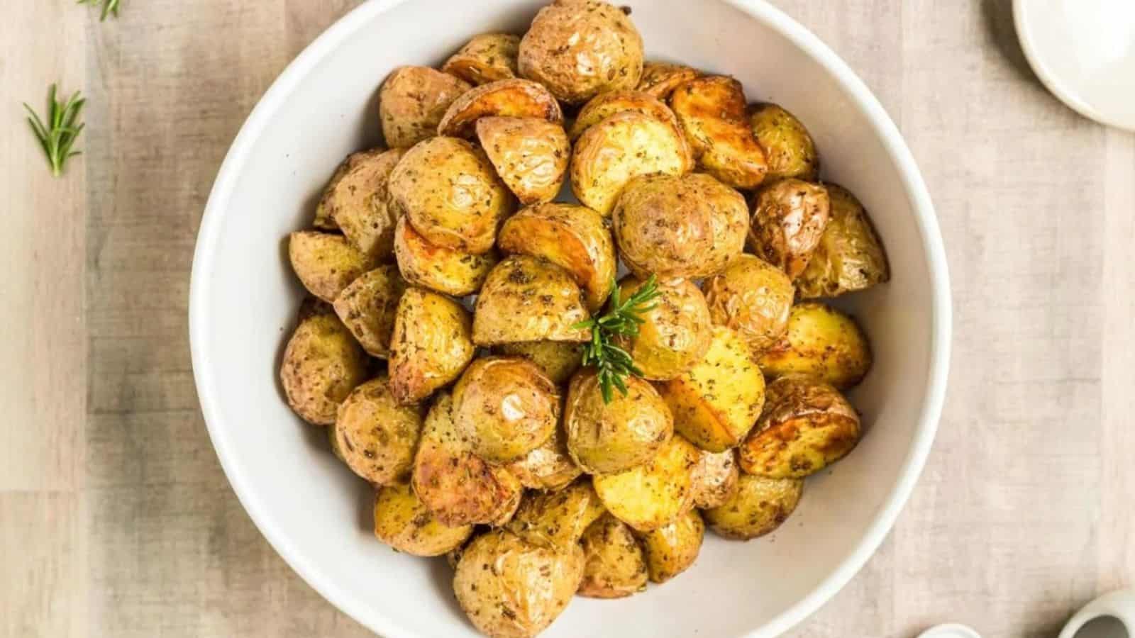 A white bowl filled with roasted potato halves and quarters, seasoned with herbs and garnished with a sprig of rosemary, sits on a light-colored wooden surface.