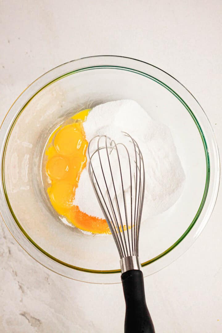 A glass bowl with eggs and sugar, and a whisk resting inside, sits on a white countertop—ingredients coming together to create a delicious Gluten Free Almond Eclair Cake.
