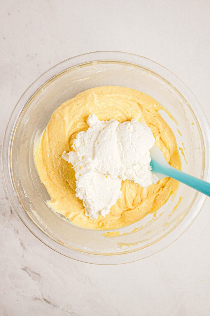 A glass bowl with yellow Gluten Free Almond Eclair Cake batter and a dollop of white cream being mixed with a blue spatula.
