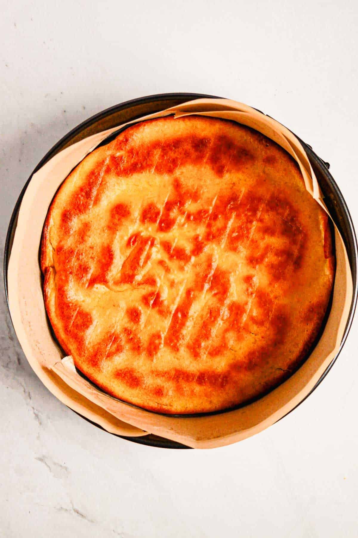 Golden-brown baked cheesecake in a round pan lined with parchment paper, reminiscent of a Gluten Free Almond Eclair Cake, viewed from above.