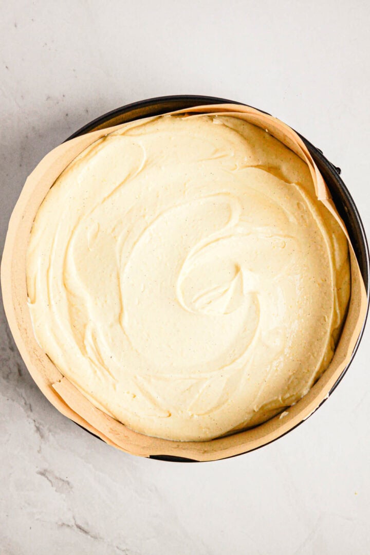Round cake pan lined with parchment paper, filled with swirled, unbaked Gluten Free Almond Eclair Cake batter on a light surface.