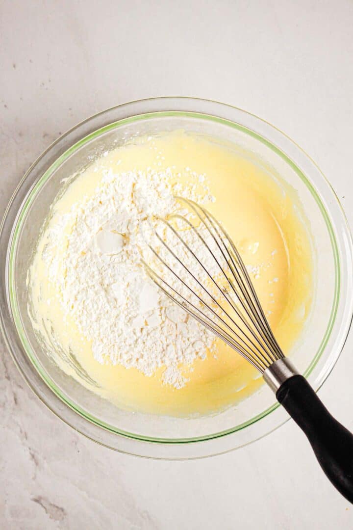 A glass bowl with batter, flour on top, and a whisk on a white countertop—perfect for preparing a Gluten Free Almond Eclair Cake.