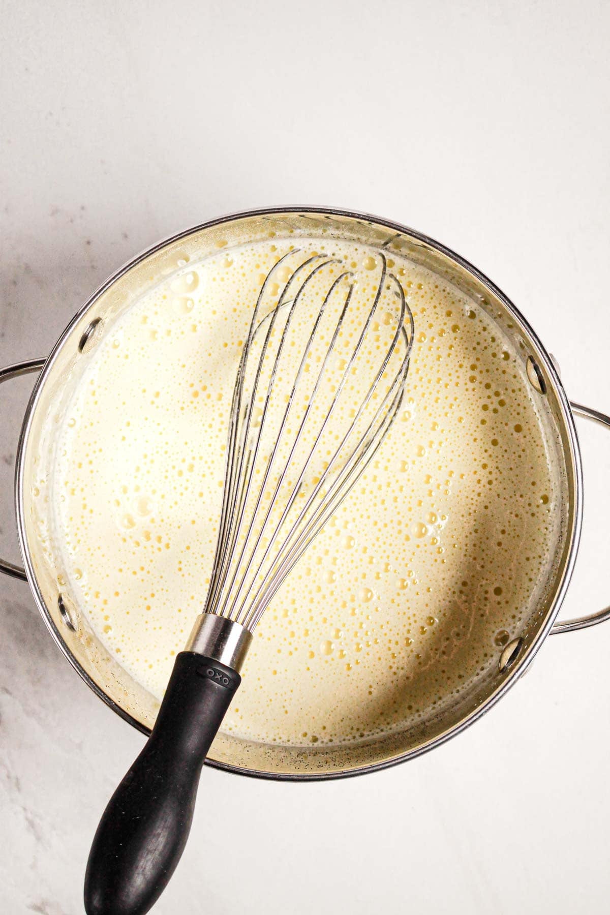 A metal pot filled with a creamy Gluten Free Almond Eclair Cake mixture and a whisk resting inside on a white surface.
