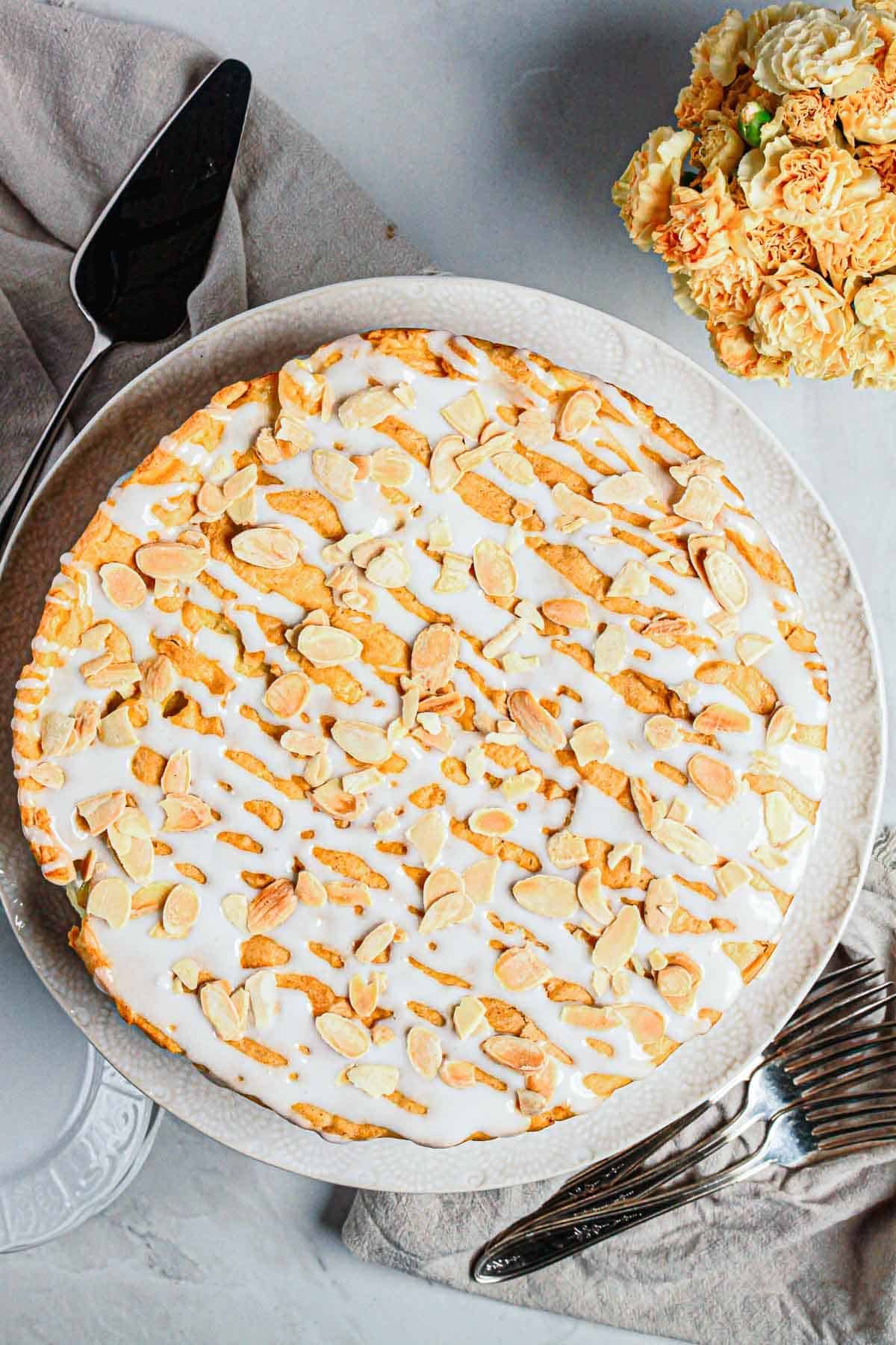 Round Gluten Free Almond Eclair Cake with white icing and sliced almonds on top, served on a white plate with utensils nearby.