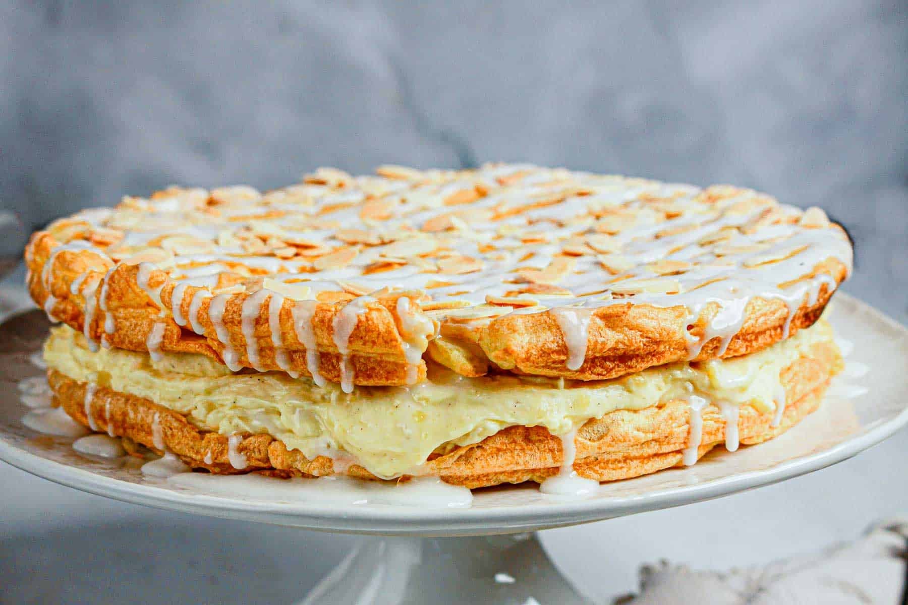 Gluten Free Almond Eclair Cake layered with cream filling, almond slices, and a white icing drizzle on a cake stand.