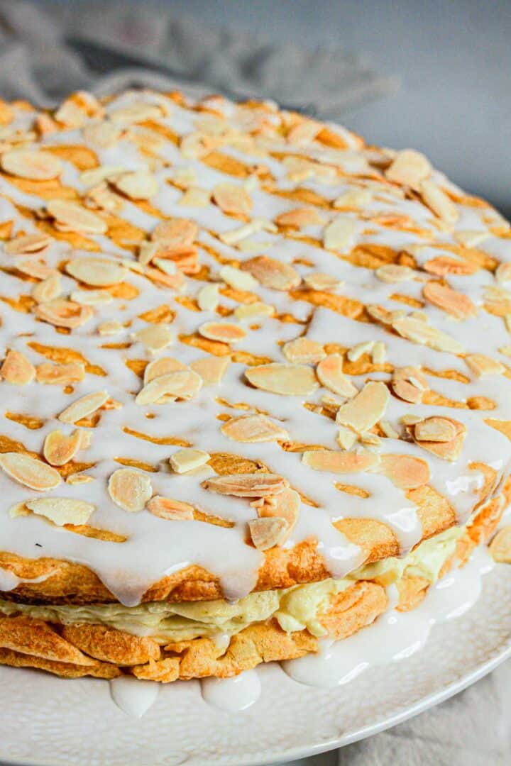 Gluten Free Almond Eclair Cake: Round cake topped with sliced almonds and white icing, with a creamy filling visible between the gluten-free cake layers.