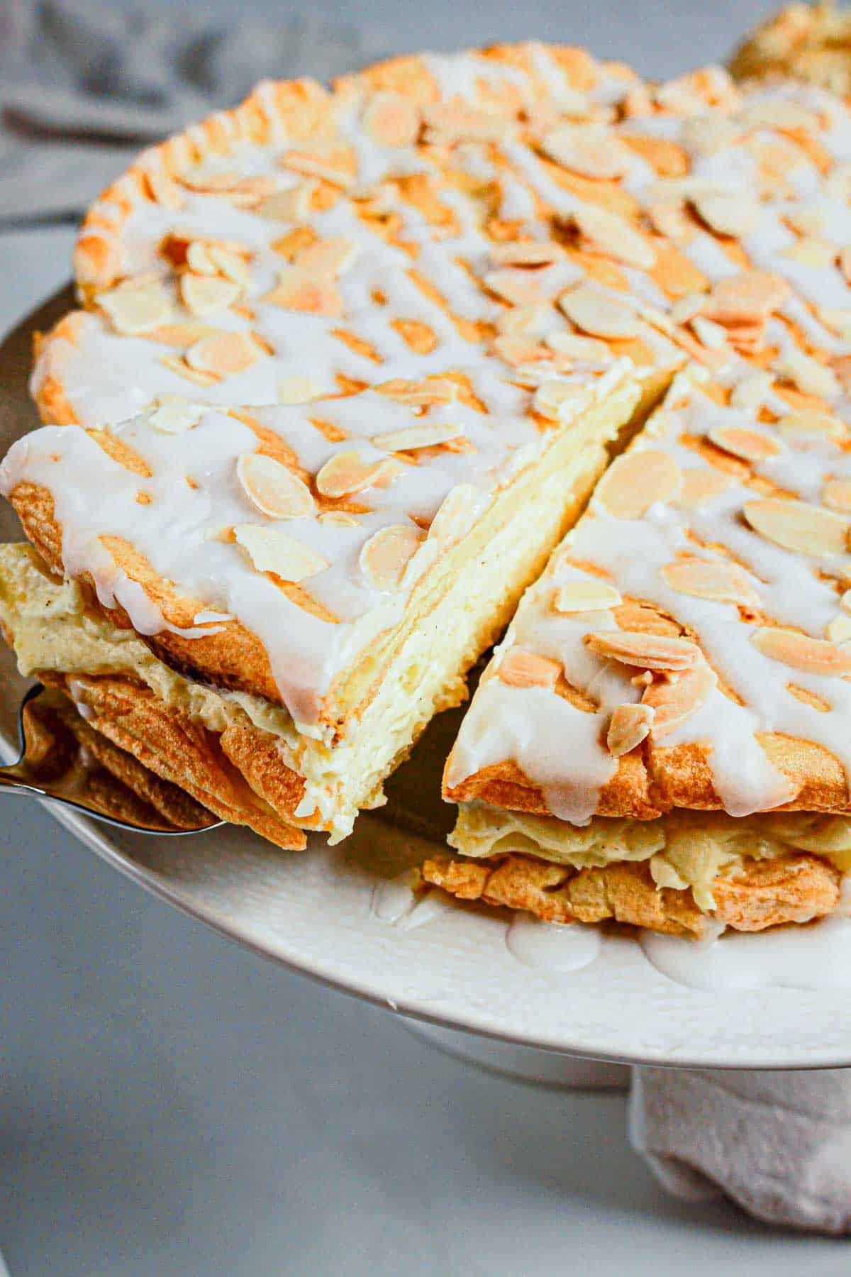 A slice of Gluten Free Almond Eclair Cake, topped with almonds and white icing, is being lifted from a plate.