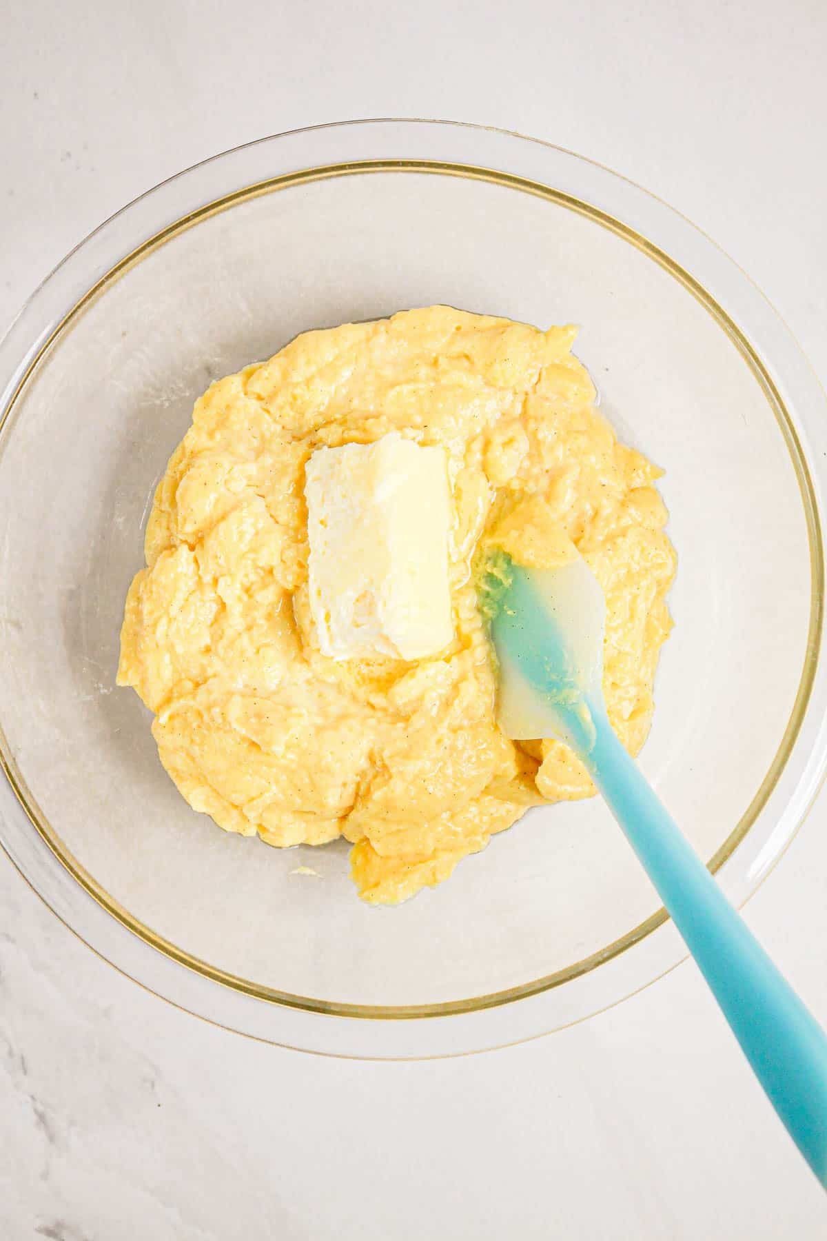 Clear glass bowl with yellow batter and a stick of butter, being mixed with a blue spatula on a white surface—perfect for preparing delicious Gluten Free Almond Eclair Cake.