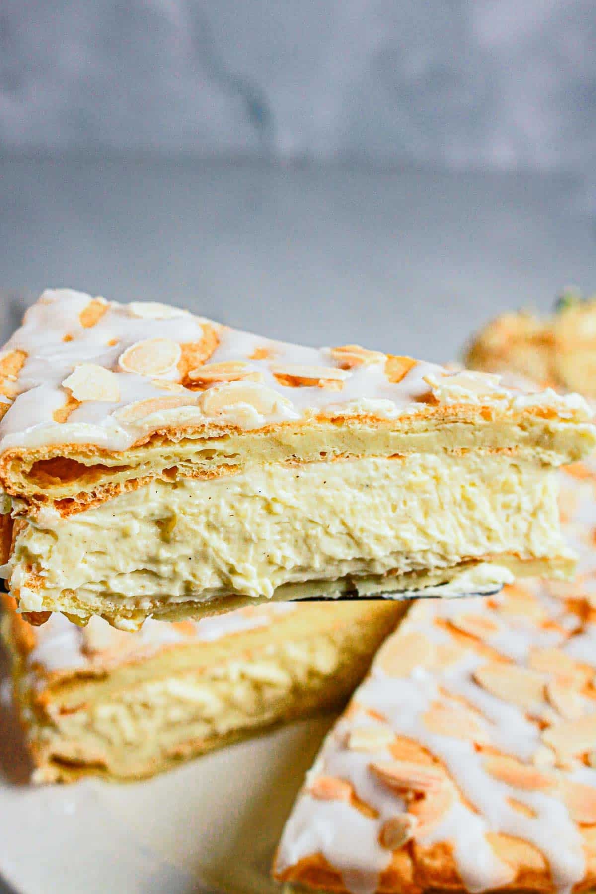 A slice of creamy, layered Gluten Free Almond Eclair Cake with icing and almond slices is being served from a whole cake.