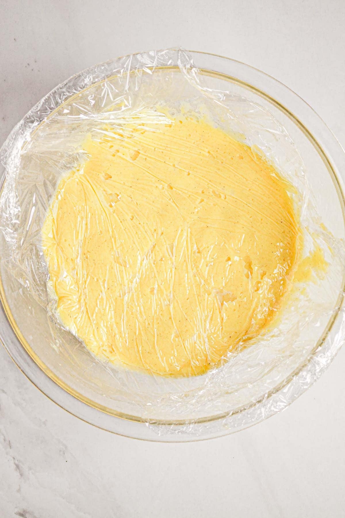A glass bowl of dough for Gluten Free Almond Eclair Cake, covered with plastic wrap and resting on a white surface.
