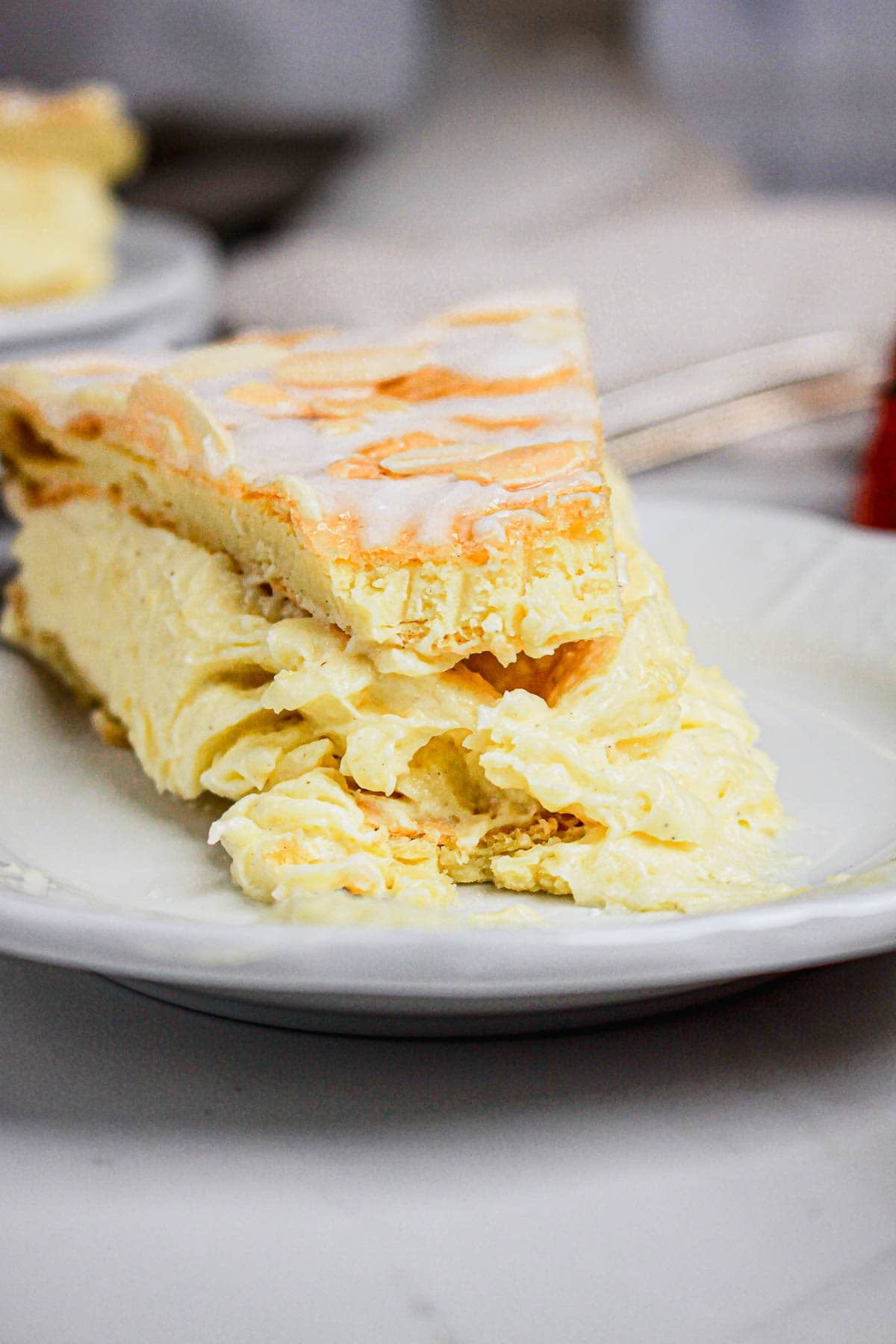 A slice of creamy, Gluten Free Almond Eclair Cake with delicate layers and a light glaze, served on a white plate.