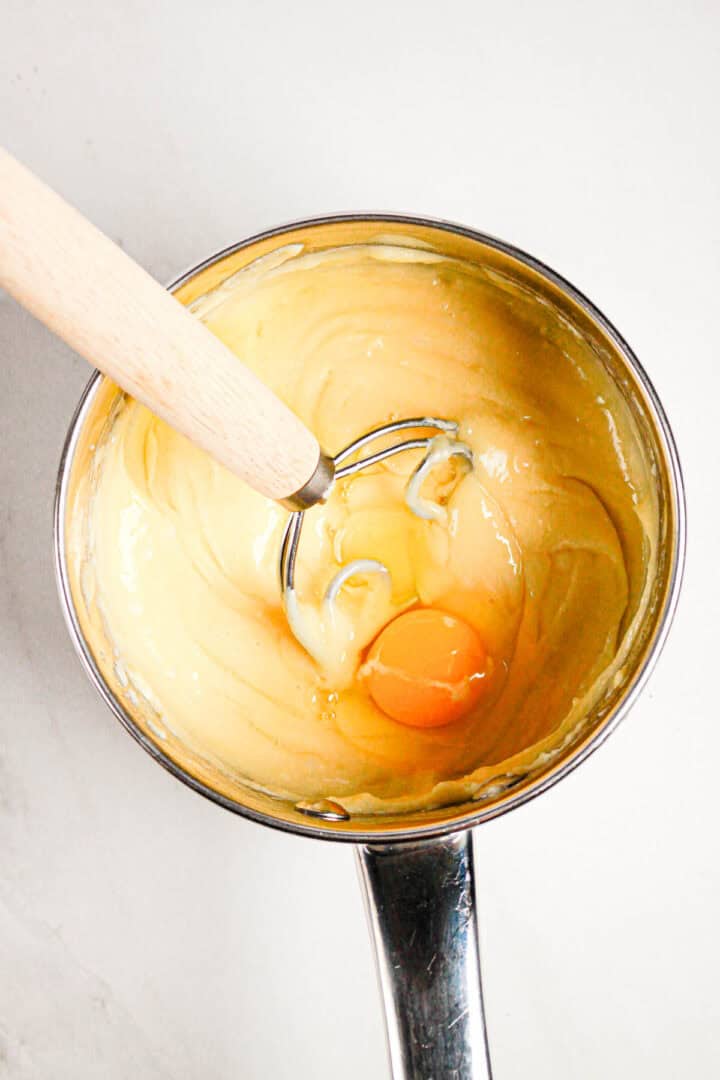 A saucepan with thick batter, a raw egg, and a whisk on a white surface—perfect for preparing Gluten Free Almond Eclair Cake.