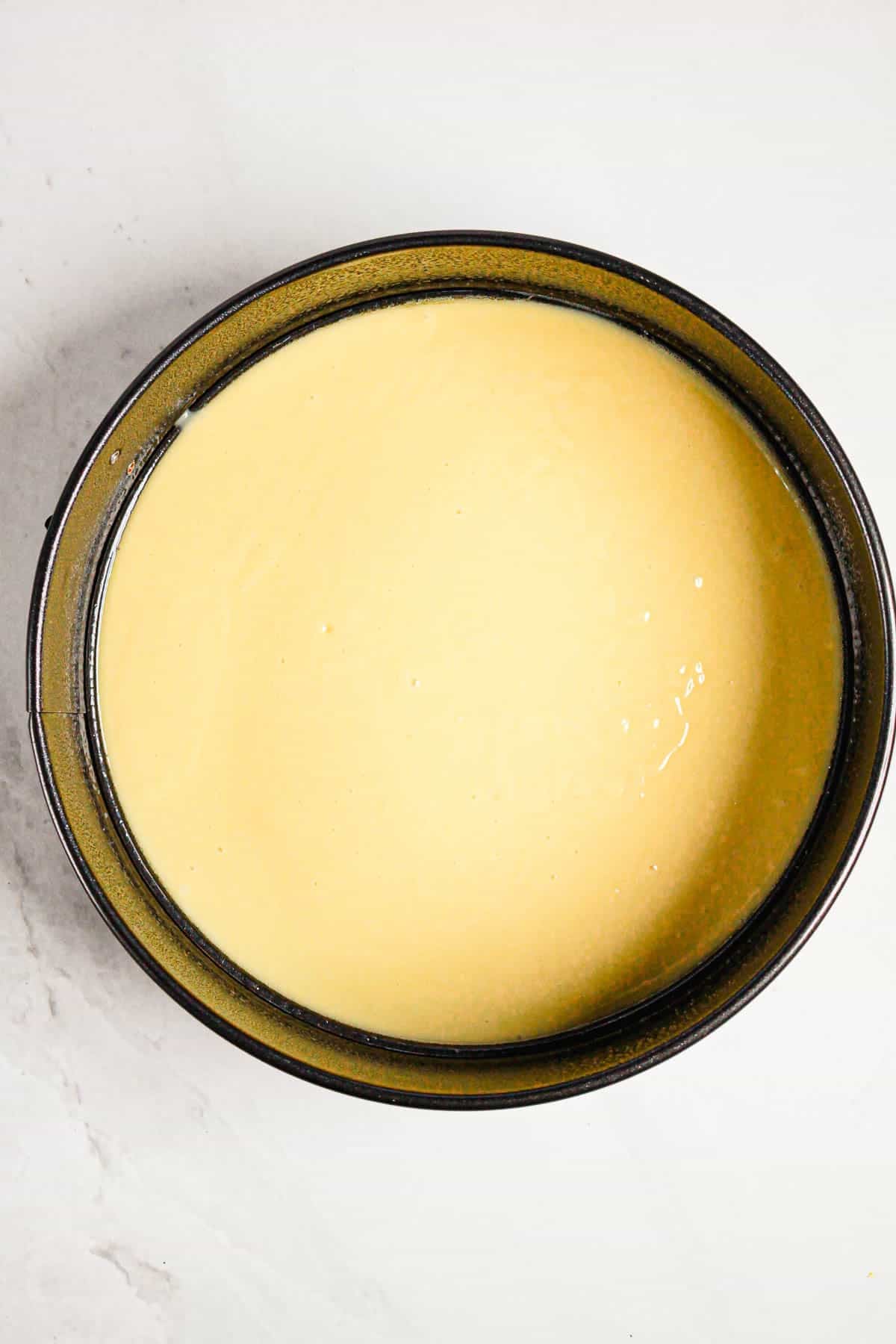 Round cake pan filled with smooth, pale yellow Gluten Free Almond Eclair Cake batter on a white surface.