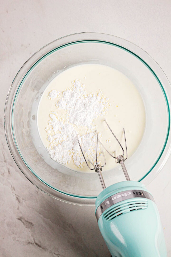 Overhead view of a glass bowl with cream and powdered sugar, and a hand mixer ready to blend for making Gluten Free Almond Eclair Cake.