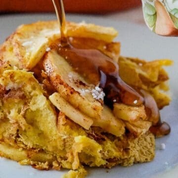 A slice of baked French toast with apples, being drizzled with syrup on a white plate.