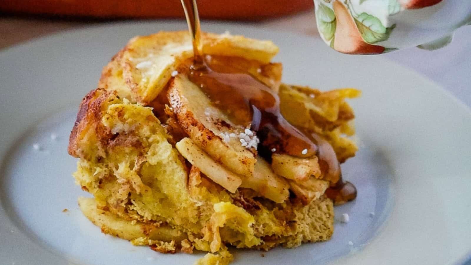 Piece of French toast casserole on a plate with a dusting of powdered sugar and a pitcher of syrup.