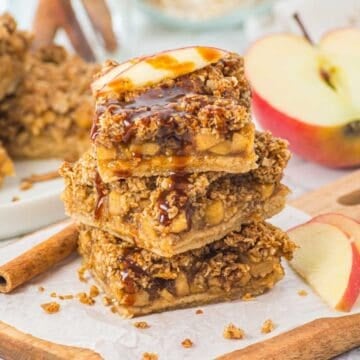 Three apple crumble bars stacked with caramel drizzle, garnished with apple slices, on parchment paper.