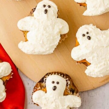 Cupcakes decorated with white frosting shaped like ghosts, displayed on a wooden board with a red napkin nearby.