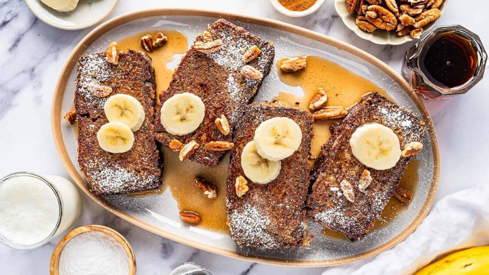 A platter of banana bread french toast with powdered sugar, banana slices, and pecans, drizzled with syrup.
