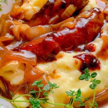 Sausages with onion gravy served over mashed potatoes, garnished with fresh thyme on a patterned plate.