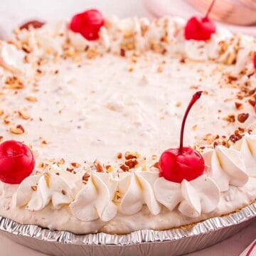 A cream pie topped with whipped cream, chopped nuts, and maraschino cherries in a foil pie tin.