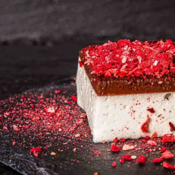 A square dessert with a white base, chocolate top layer, and red berry pieces sprinkled on top and around.