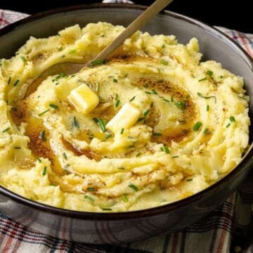 Bowl of mashed potatoes topped with butter, chives, and pepper on a plaid cloth.