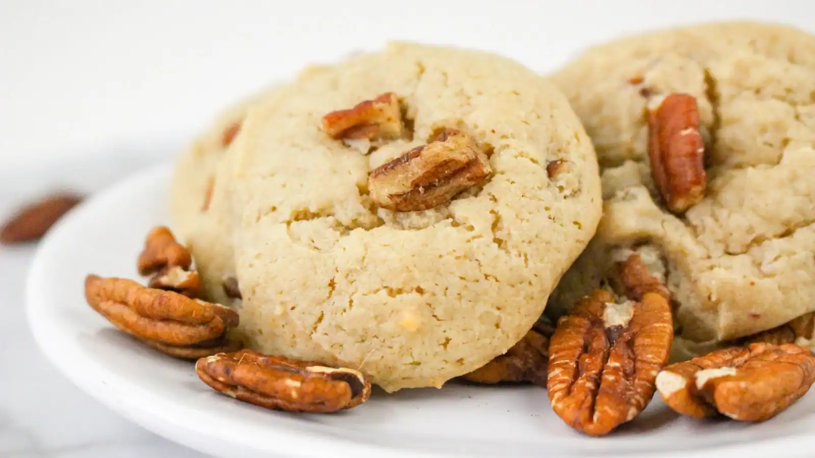 Two pecan cookies on a white plate, surrounded by whole pecan nuts.