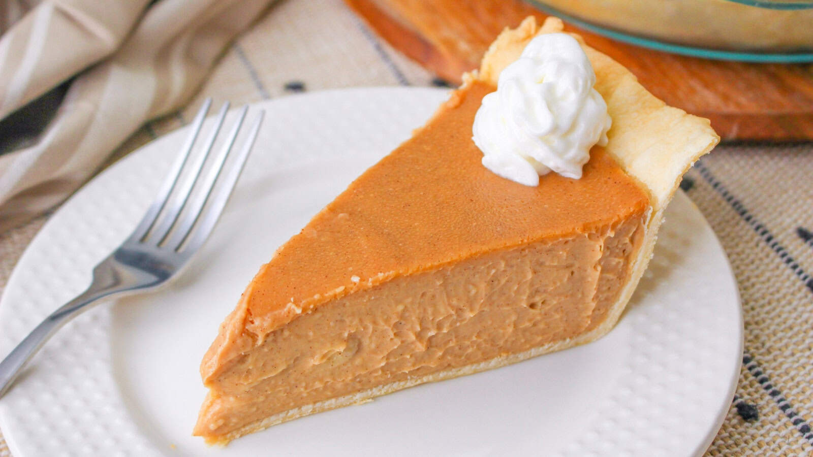 A slice of butterscotch cinnamon pie topped with whipped cream on a white plate, accompanied by a fork on a linen surface.