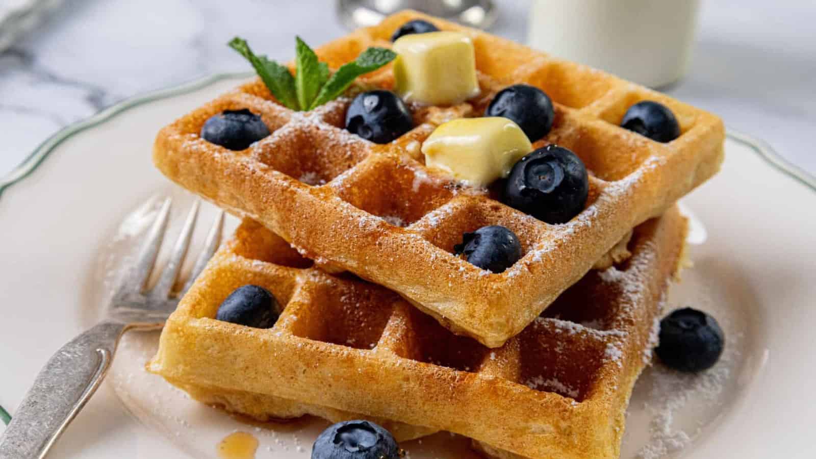 Close-up of golden brown waffles topped with blueberries, butter cubes, a sprinkle of powdered sugar, and a small mint leaf. A fork is partially visible in the foreground.