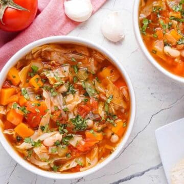 Two bowls of vegetable soup with beans, carrots, and herbs, next to bread slices, tomato, and cilantro.