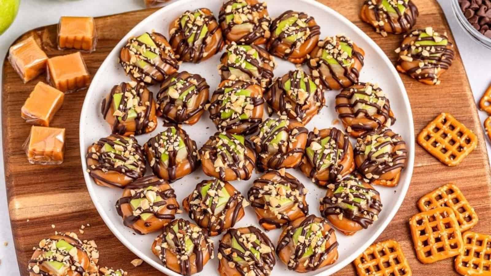 Caramel apple pretzel bites topped with chocolate drizzle and chopped nuts on a serving plate.