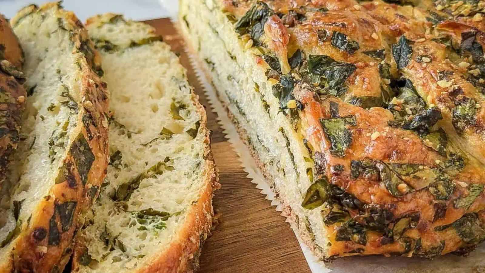 Close-up of a sliced, freshly baked herb pull-apart bread on a wooden cutting board. The bread's crust is golden brown and speckled with finely chopped herbs. The interior appears soft and fluffy, with visible green herbs swirled throughout the bread.