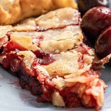 A slice of cherry pie with flaky crust on a plate, served with whole cherries.
