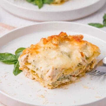 A slice of cheesy lasagna on a white plate with a fork and basil garnish.