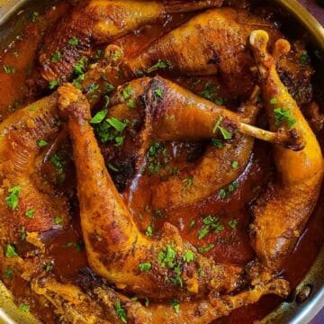 Chicken legs cooked in a rich red sauce, garnished with chopped herbs in a pan on a wooden table.
