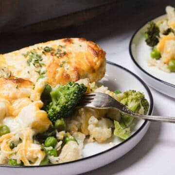 A plate of cheesy vegetable casserole with broccoli, peas, and cauliflower, with a fork lifting a bite.